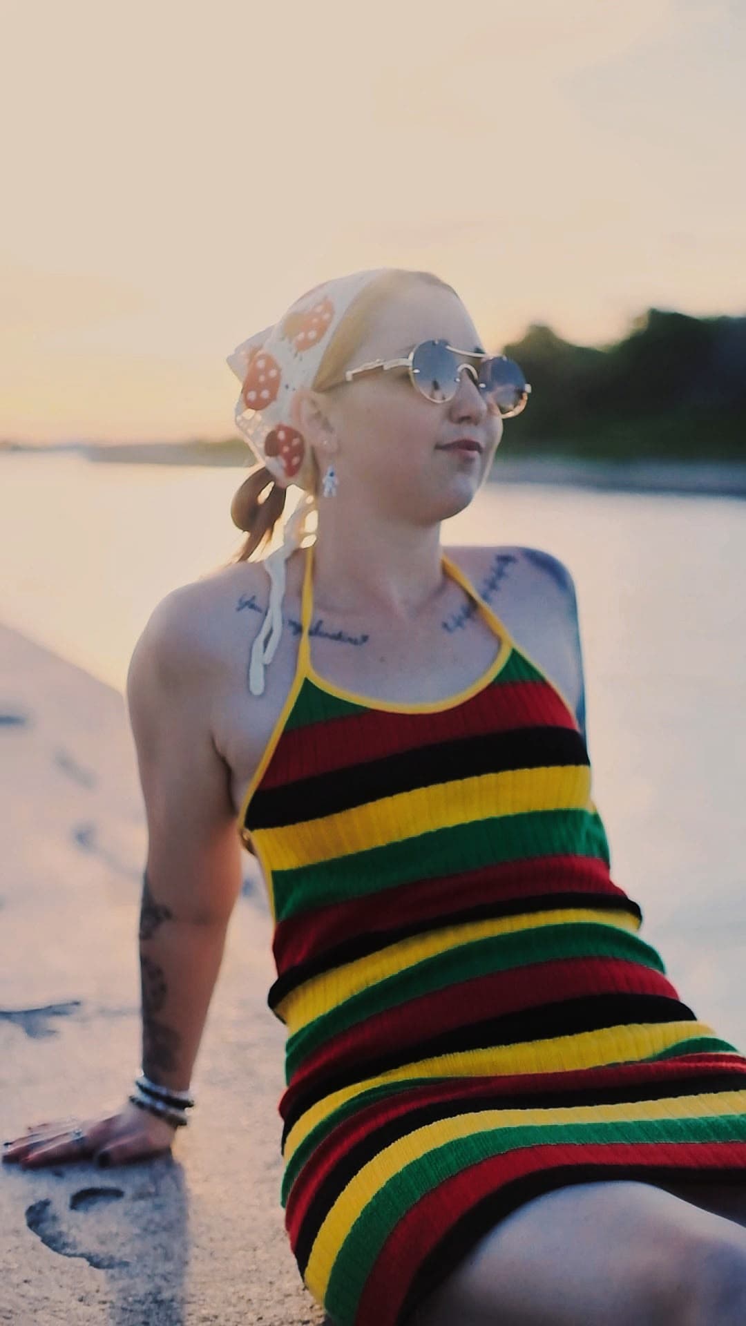 A woman in a bandana and round sunglasses relaxing on a seawall by the water at sunset
